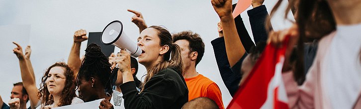 Mehrere Personen demonstrieren. Eine Person ind er Mitte spricht in ein Megafon, andere Personen halten Flaggen und Transparente.