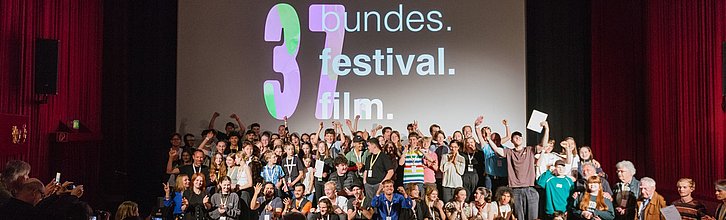 Eine sehr große Gruppe von Menschen steht auf einer Bühne. Auf der Leinwand im Hintergrund steht 37. bundes.festival.film