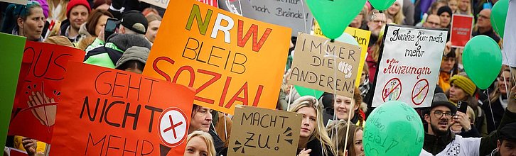 Bild von NRW Bleib sozial Demo mit Demo-Teilnehmenden und Plakaten. Im Fokus ein Plakat mit der Aufschrift "NRW bleib sozial"