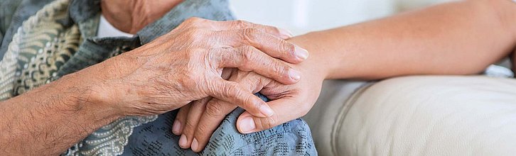 Eine ältere Person hält die Hand einer jüngeren Person, die dieser ihr auf die Schulter gelegt hat.