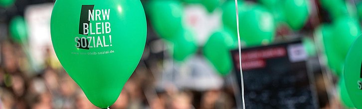 Ein grüner Lufballon mit dem Aufdruck "NRW bleib sozial". Im Hintergrund sind noch mehr Ballons und eine demonstrierende Menge zu sehen.