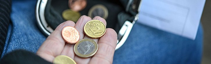 Eine offene Handfläche, auf der mehrere Euro-Münzen liegen, über einer schwarzen Geldbörse. Im Hintergrund liegt eine Rechnung.