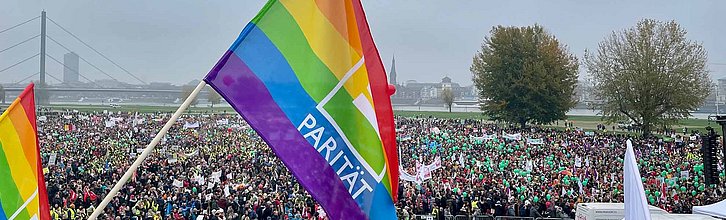 Eine Regenbogenflagge mit dem Schriftzug Parität im Vordergrund, im Hintergrund eine Menschenmenge bei einer Demo