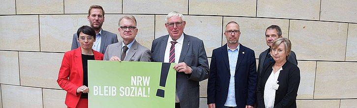 Sieben Personen stehen nebeneinander und halten ein Schild mit der Aufschrift "NRW bleib sozial!" hoch