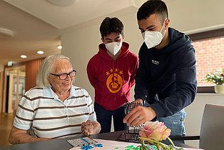 Zwei männliche Jugendliche tragen FFP2-Masken und hefen einer Seniorin beim Bingo-Spiel.