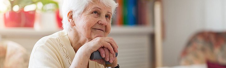 Eine Seniorin stützt ihre Hände und ihr Kinn auf ihre Gehhilfe mit einem erleichterten Gesichtsausdruck.